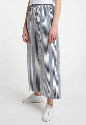 Personne portant un pantalon large à rayures verticales blanches et bleu marine avec une taille élastique et des baskets blanches, debout devant un fond uni.