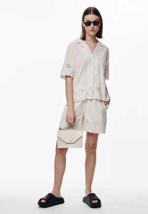 Lys beige to-deltant antrekk som inkluderer en kortermet, knappet skjorte og matchende shorts med sikksakkkanter og hullmønster. Kombinert med en teksturert clutch og svarte sandaler.