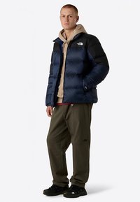 Navy blue insulated puffer jacket with black shoulders, zipper pockets, and a high collar, paired with olive green pants and black sneakers.