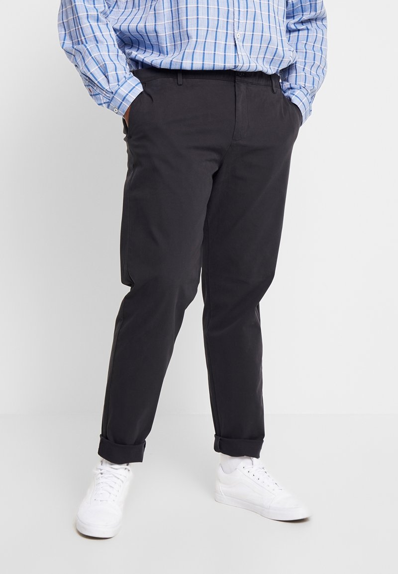 Pantalons en coton noirs à coupe slim, avec des ourlets retroussés et des poches latérales, associés à des baskets blanches et une chemise à carreaux bleue.