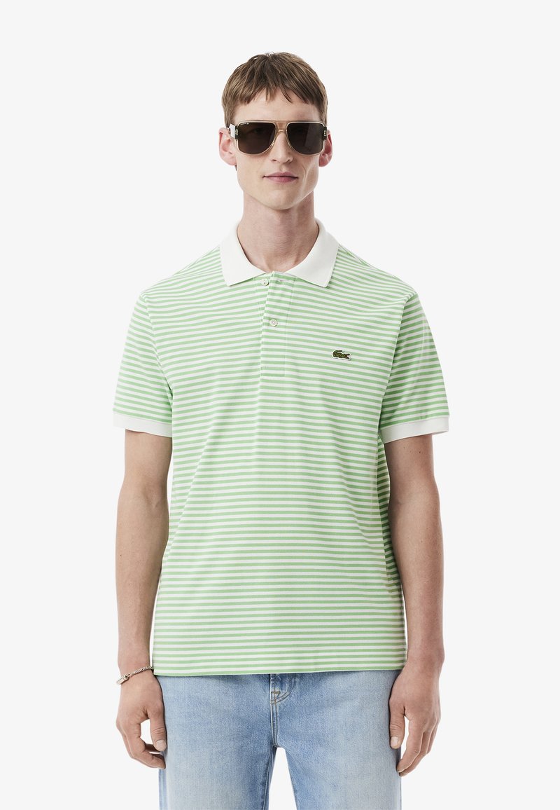 Grön- och vitrandig polo med krage och korta ärmar, med en liten broderad logotyp på bröstet. Bärs med solglasögon.