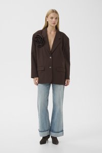 Brązowa, oversize'owa marynarka z dużym kwiatowym detalem na ramieniu, w komplecie z jasnoniebieskimi, szerokimi dżinsami i czarnymi botkami na obcasie.