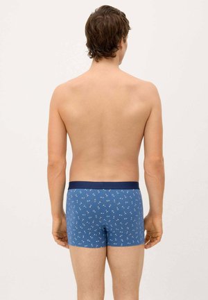 Hombre de pie con la espalda hacia la cámara, vistiendo bóxer azul con un pequeño patrón floral y cintura azul marino.