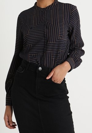 Femme portant un chemisier noir boutonné avec des lignes géométriques beiges, rentré dans une jupe noire taille haute, main reposant dans la poche de la jupe.