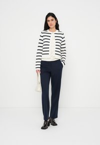 Λευκό ζακέτα με ναυτικές ρίγες, κλείσιμο με κουμπιά, σετ με ναυτικό παντελόνι και μαύρα παπούτσια. Η τσάντα είναι ανοιχτό γκρι, προσθέτοντας αντίθεση.