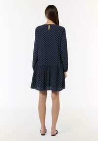 Navyblaues Kleid mit langen Ärmeln, einem ausgestellten Saum und einem Schlüsselloch-Detail im Rücken. Mit einem geometrischen weißen Muster throughout dekoriert.