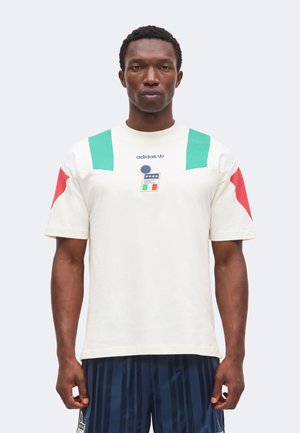 Άνδρας που φοράει λευκό T-shirt Adidas με πράσινα και κόκκινα γεωμετρικά σχήματα και λογότυπο, συνδυασμένο με σκούρο μπλε ριγωτή βερμούδα, στέκεται μπροστά σε γκρι φόντο.