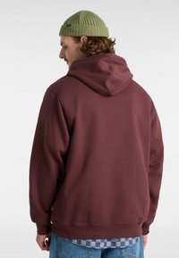 Maroon hoodie med huva och kenguruficka, tillverkad av mjukt material. Matcher med en blå rutig skjorta under. Grön ribbad mössa på huvudet.