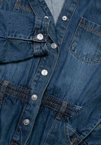 Džínová bunda s kovovými patentkami, náprsní kapsou a elastickým pasem, vykazující detailní prošívání a ohrnuté manžety.