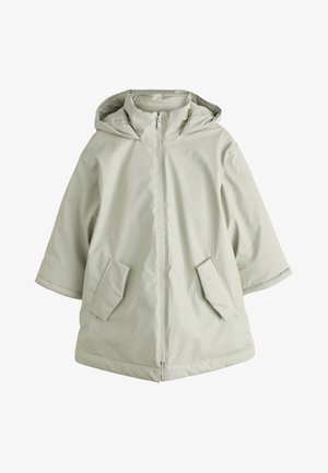 Unge barn iført en lysegrøn hættetrøje, rød cardigan med knapper, stribet skjorte, jeans med vide ben og lyserøde sneakers, der smiler.