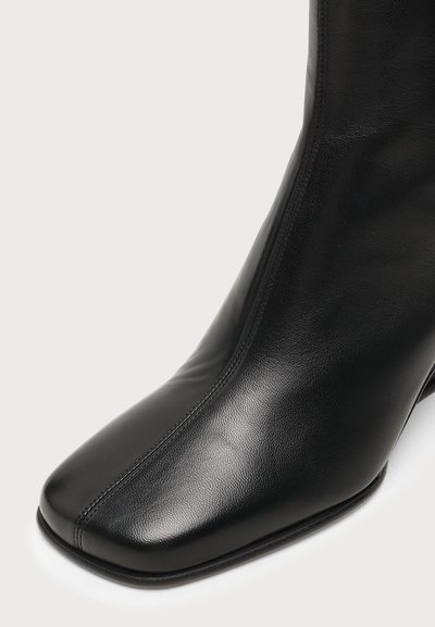 Bottine noire en cuir avec bout carré et couture apparente le long de la couture centrale avant sur fond blanc.