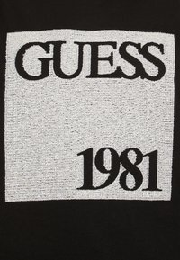 Svart klädsel med en strukturerad silverrektangulär lapp där varumärket "GUESS" står över och "1981" under i fet svart stil.