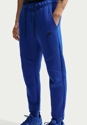 Mand iført blå Nike joggers og matchende blå sneakers, stående med den ene hånd i lommen og synlig tatovering på underarmen.