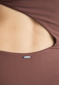 Brun uten ermer topp med utskjæringsdesign, glatt tekstur og en metallisk merkeetikett festet på siden.