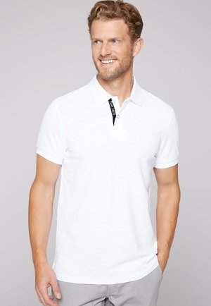 Camisa polo blanca de manga corta, con un acento en el cuello negro y un sutil logotipo de la marca bordado en el pecho. Textura de tela suave.