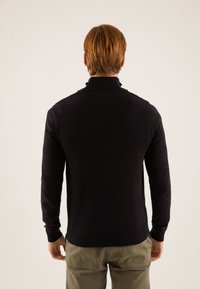 Černý svetr s vysokým límcem, texturovaným pleteným vzorem, dlouhými rukávy a žebrovanými manžetami, nošený s světlými kalhotami.