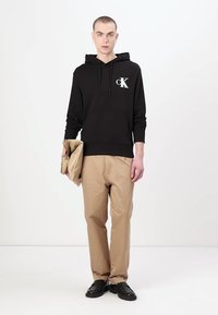 Felpa nera in cotone con tasca frontale e logo, abbinata a pantaloni color sabbia. L'outfit è completato da scarpe nere, con una borsa in mano.