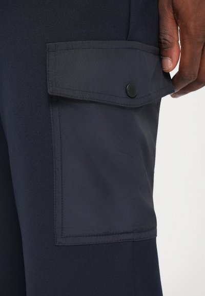 Pantaloni cargo bleumarin, cu un buzunar lateral cu clapetă și închidere prin capse, fabricați dintr-un material texturat cu detalii vizibile de cusătură.