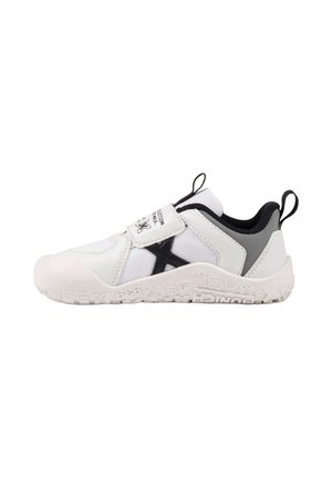 Zapatilla deportiva blanca y negra con tira de velcro, suela texturizada y lengüetas en el talón y la lengüeta, diseñada para fácil uso y comodidad.