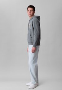 Felpa grigia con cappuccio e tasca frontale, con design di testo bianco, abbinata a jeans azzurri e sneakers bianche. Outfit semplice e casual.