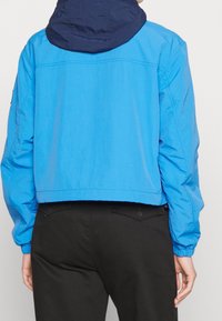 Cropped blue jacket with a navy hood. Features long sleeves and elastic cuffs. Smooth texture and minimal design with no visible logo.