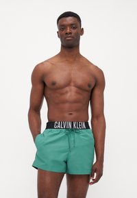Zelene kupaćice s elastičnom crnom pasicom koja sadrži logo "CALVIN KLEIN". Glatka tekstura, kroja iznad koljena i detalj svezanja.