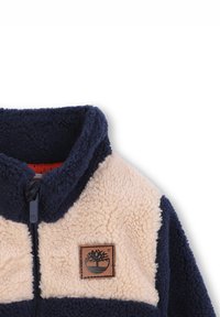 Giacca in pile blu navy e beige con colletto rialzato, zip frontale e una toppa con logo marrone sul lato sinistro del petto.