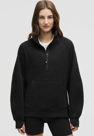 Junge Frau, die einen schwarzen Reißverschluss-Sweatshirt mit Kängurutasche und schwarzen Shorts trägt, steht vor einem schlichten grauen Hintergrund.