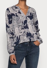 Donna che indossa una blouse blu navy con un motivo paisley bianco e jeans blu scuro, con una mano in tasca, su uno sfondo neutro.