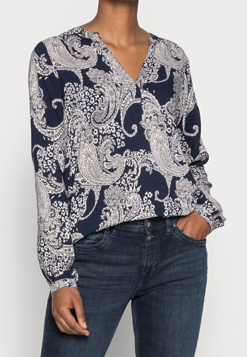 Donna che indossa una blouse blu navy con un motivo paisley bianco e jeans blu scuro, con una mano in tasca, su uno sfondo neutro.