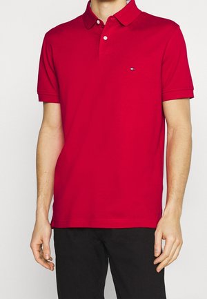 Mand iført en rød polo t-shirt med korte ærmer og et lille logo på venstre bryst samt sorte bukser, stående mod en ensfarvet baggrund.