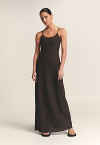 Maxikleid aus dunkelbraunem Stoff, Spaghettiträger, figurbetontes Oberteil und fließender Rock. Kombiniert mit schwarzen und beigen Sandalen, minimalistisch gestaltet.