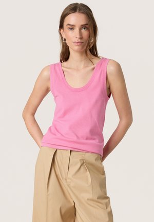 Femme portant un top rose sans manches et un pantalon beige plissé à jambes larges, posant avec les mains dans le dos sur un fond uni.