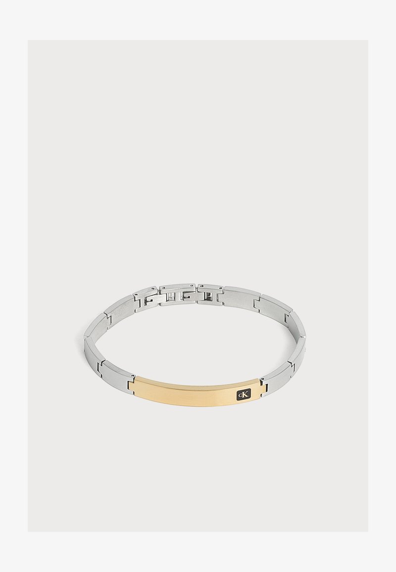 Bracelet à maillons en métal argenté avec une plaque rectangulaire dorée ornée d’un petit logo Calvin Klein noir sur un fond uni clair.
