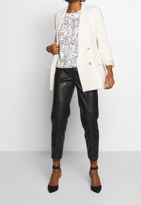Blazer bianco doppiopetto indossato su un top a motivi bianco e nero con design astratti, abbinato a pantaloni in pelle nera a gamba affusolata e tacchi neri.