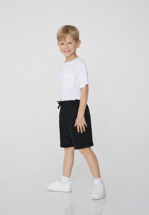 T-shirt bianco in cotone con maniche corte e taschino sul petto, abbinato a pantaloni corti neri con coulisse. Indossato con sneaker bianche e calzini bianchi.