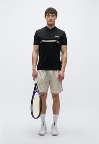 Svart poloshirt med korte ermer, en glidelås og horisontale striper, kombinert med lyse shorts og en tennisracket.