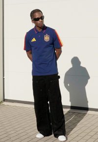 Laivastonsininen poolopaita, jossa punaisia ja keltaisia yksityiskohtia, mustat leveälahkeiset samettihousut, valkoiset sukat ja aurinkolasit. Seisoo vaaleaa taustaa vasten.