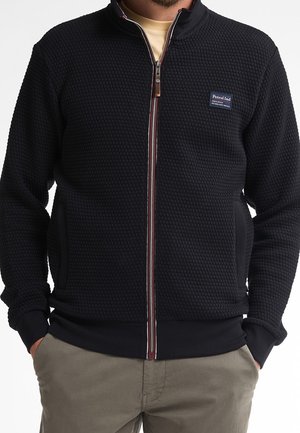 Man draagt een gestructureerde zwarte jas met rits en een klein "Petrol Ind" logo op de borst, handen in de zakken, gecombineerd met beige broek.