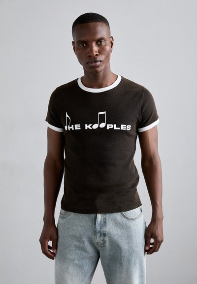 T-shirt marron foncé avec un col et des manches blanches, affichant le texte "THE KOOPLES" et des notes de musique imprimées en blanc.