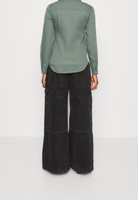 Pidempi oliivinvihreä paita, jossa on kaareva helma, yhdistettynä leveälahkeisiin mustiin cargo-housuihin, joissa on sivutaskut ja vetoketjut.