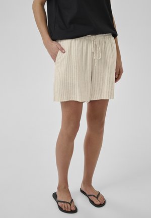 Frau trägt beige Shorts mit Kordelzug und Taschen mit Nadelstreifen, schwarze Flip-Flops und ein schwarzes Top, steht vor einem schlichten Hintergrund.