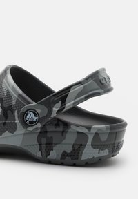 Sabots à imprimé camouflage dans des nuances de gris et noir, fabriqués en matériau flexible avec une tige texturée et une sangle ajustable ; présente un détail de logo.