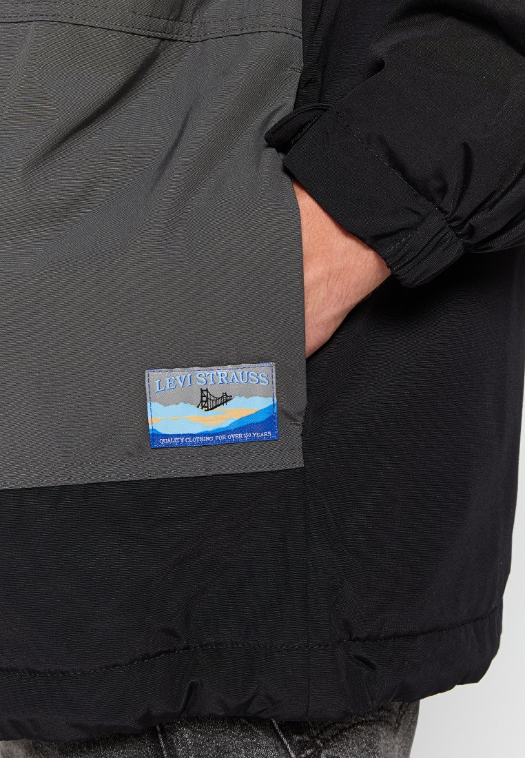 Chaqueta negra y gris con una textura de tela suave, puños elásticos y un parche rectangular del logo de Levi Strauss con detalles en azul y blanco.