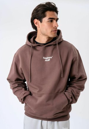 Homme portant un sweat à capuche marron avec le texte « Toujours Actif » sur le devant, mains dans les poches, regardant vers la droite sur fond blanc.