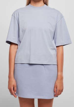 Femme portant un t-shirt ample gris clair à manches courtes et une jupe courte assortie, debout devant un fond gris uni.