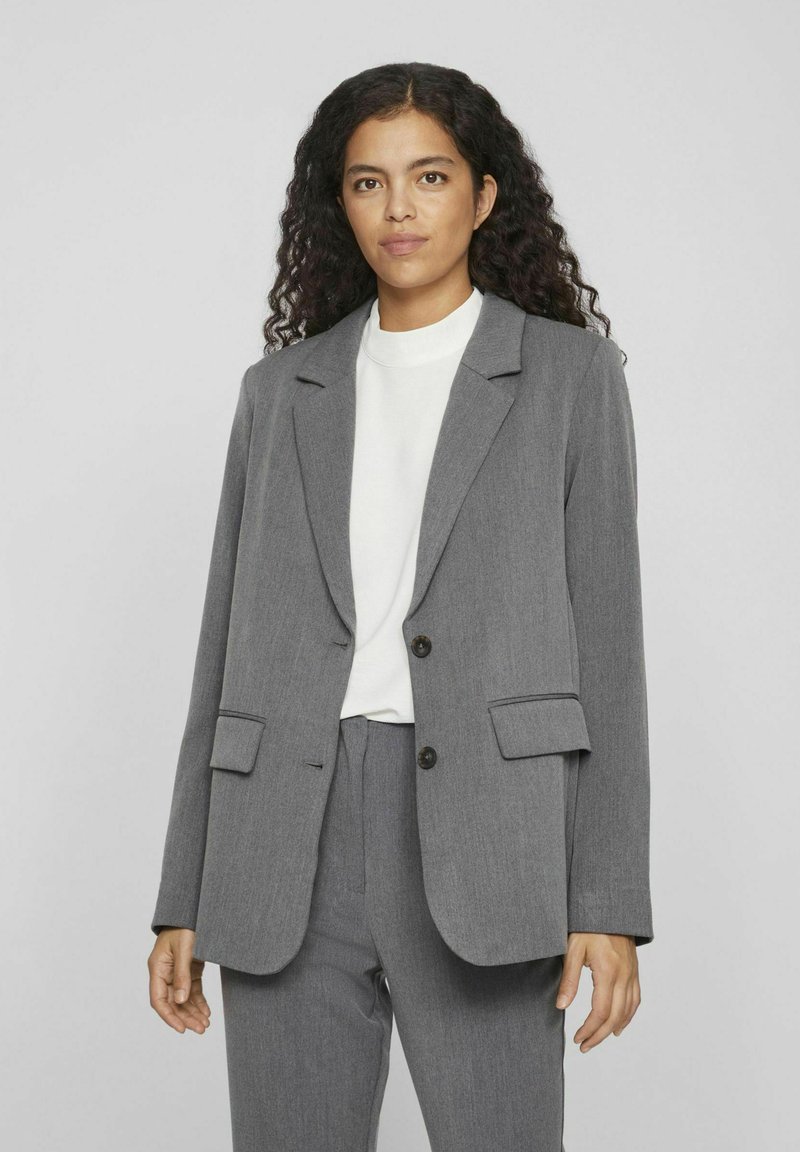 Blazer gris con solapa de muesca, dos bolsillos frontales y cierre de botón único. Hecho de tejido texturizado, combinado con una blusa blanca.