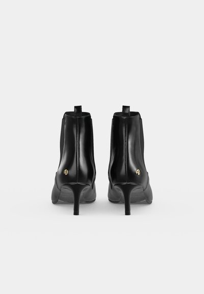 Une paire de bottines noires en cuir à talons hauts et fins, vues de dos, chacune ornée d’un petit emblème doré sur le côté.