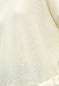 Tissu tricoté couleur crème avec une texture subtile et une petite signature brodée claire près du coin inférieur droit.