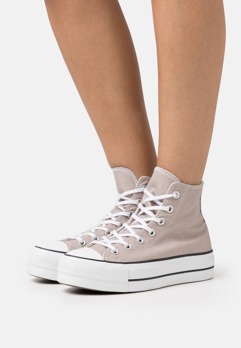 High-top sneakers in light beige canvas with white laces and a rubber sole, featuring a contrasting black stripe along the base.